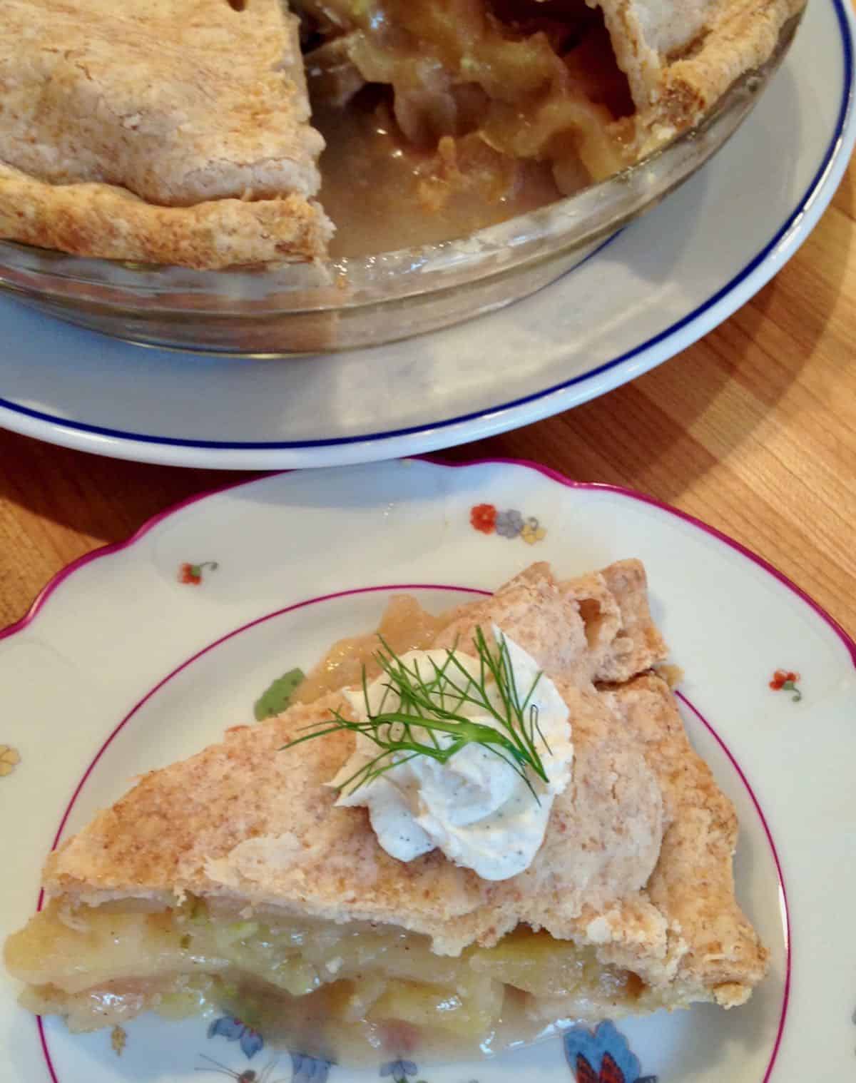 Slice of apple fennel pie topped with whipped cream on a plate with whole pie in background.