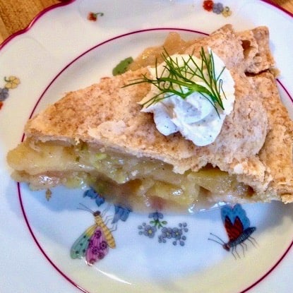 Slice of apple fennel pie topped with cream and fennel frond on a plate.