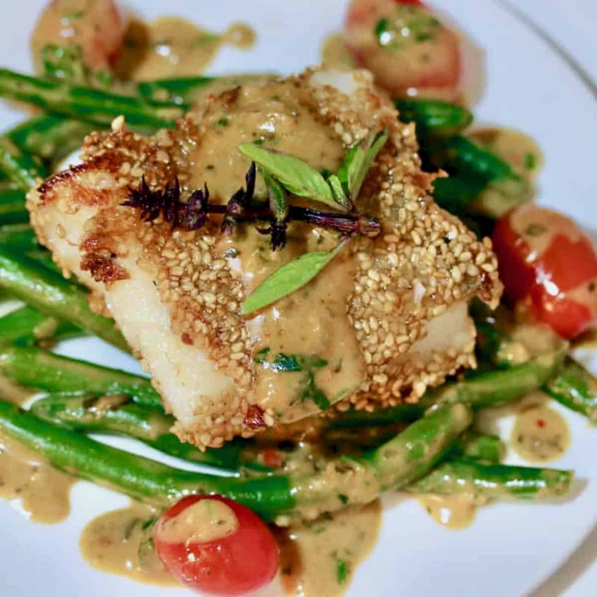 Roasted sesame cod on a plate with green beans, cherry tomatoes and lemon grass basil sauce.