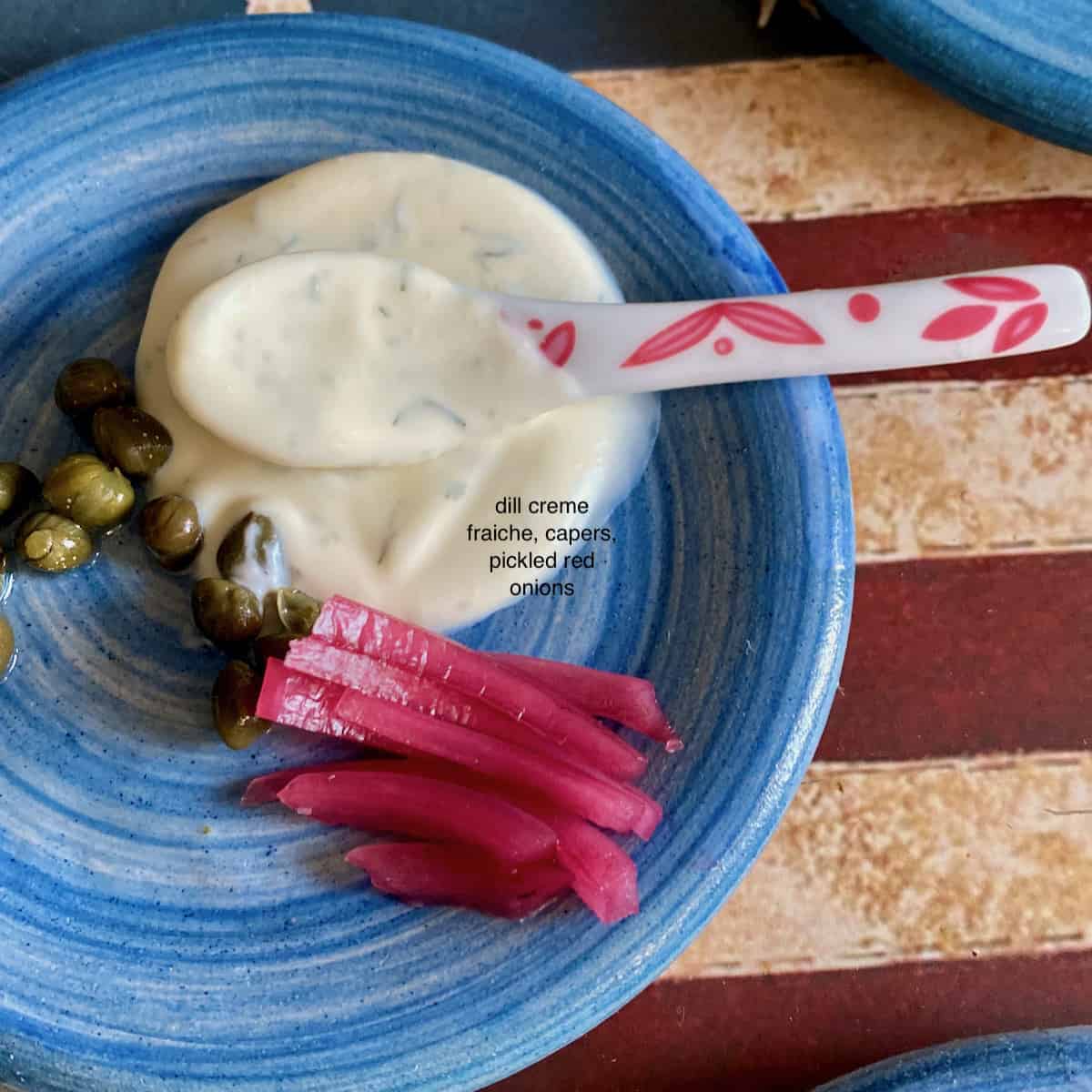 Capers, pickled red onions and creme fraiche on a blue plate with a spoon.