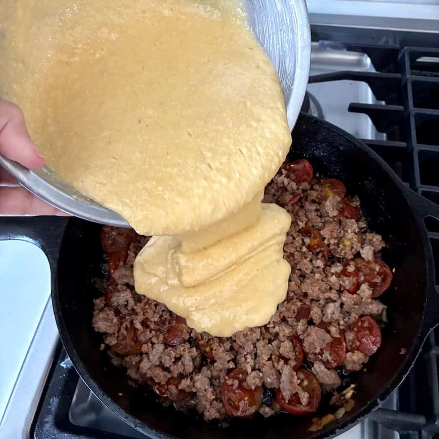 Pouring cornbread batter into a skillet with cooked sausage, onions and tomatoes.